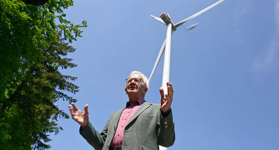 Ministerpräsident Winfried Kretschmann steht bei einer Besichtigung einer Baustelle einer Windkraftanlage vor einem schon gebauten Windrad.