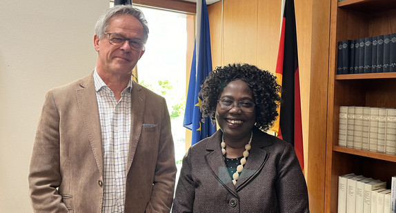 Staatssekretär Rudi Hoogvliet und I.E. Frau Annonciata Sendazirasa, Botschafterin der Republik Burundi stehen nebeneinander im Kaminzimmer der Landesvertretung und blicken in die Kamera. Im Hintergrund sieht man die europäische und die deutsche Flagge.