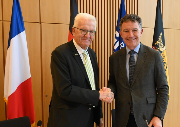 Ministerpräsident Winfried Kretschmann (links) und Franck Leroy (rechts), Präsident der Region Grand Est, stehen vor Fahnen und geben sich die Hand.