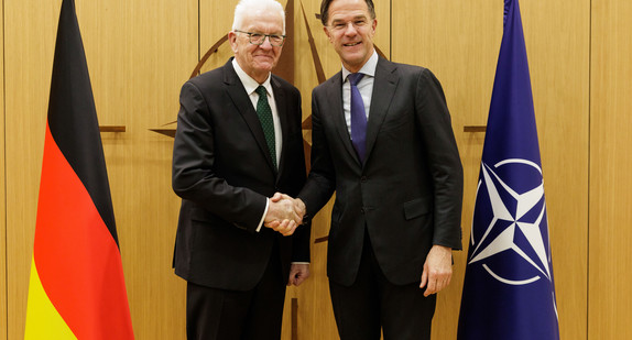 Ministerpräsident Winfried Kretschmann (links) und NATO-Generalsekretär Mark Rutte (rechts) stehen zwischen Fahnen und geben sich die Hand.