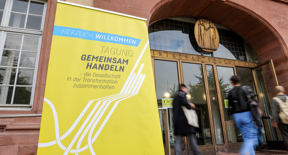 Eingang zur Tagung in der Universität Mannheim mit einem Banner mit der Aufschrift „HERZLICH WILLKOMMEN - TAGUNG GEMEINSAM HANDELN – die Gesellschaft in der Transformation zusammenhalten“.  