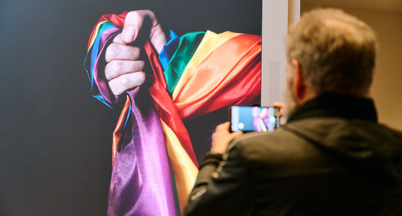 Einladungsbild der Veranstaltung auf der LED-Wand im Foyer der Landesvertretung. Ein Mann fotografiert die Hand, die eine Regenbogenfahne umschlungen hält