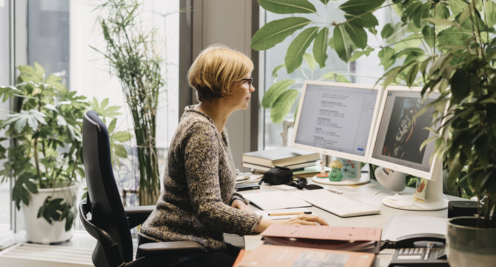 Person mit kurzem Haar und Brille sitzt an einem Schreibtisch mit zwei Monitoren, umgeben von mehreren Pflanzen und Aktenstapeln.