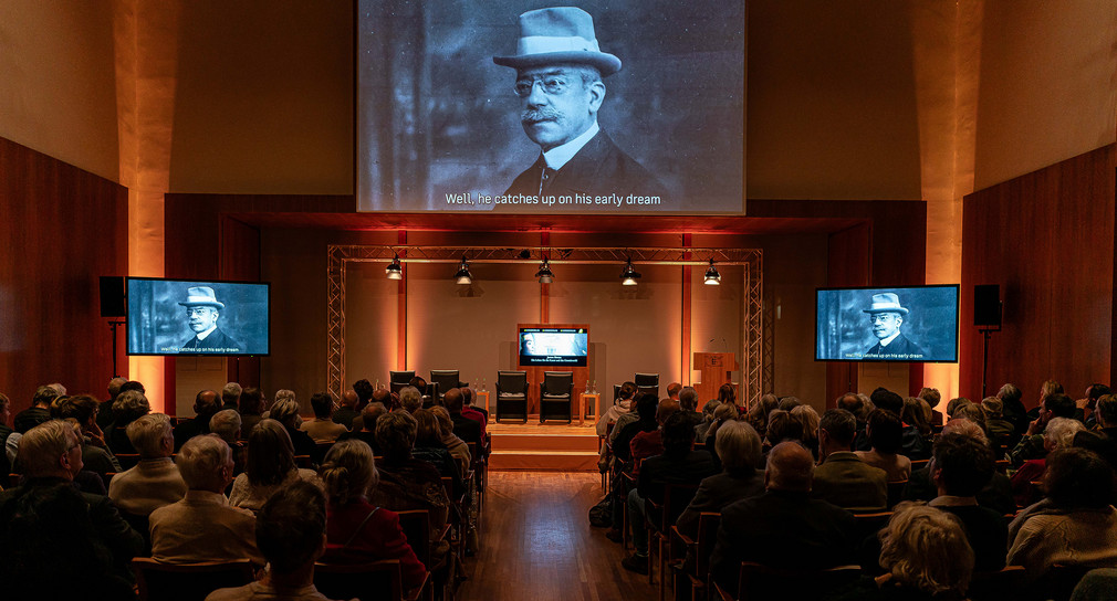 Die Veranstaltungsgäste sehen ein kurzes Video zur Biographie James Simons über die Leinwand im Großen Saal