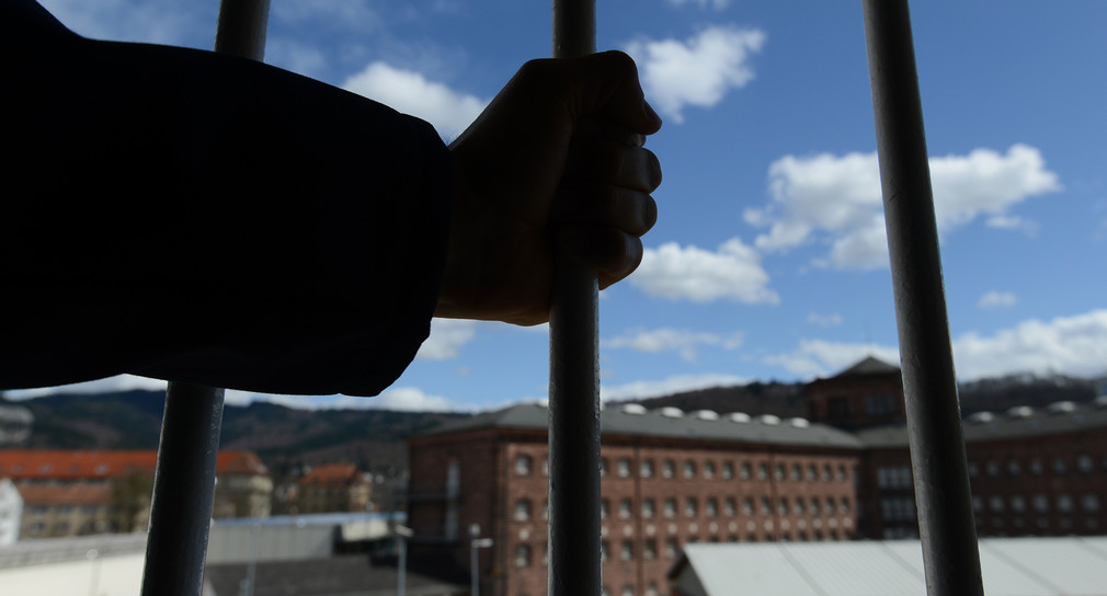 Mann greift mit seiner Hand nach den Gitterstäben eines Fensters. Im Hintergrund stehen Wohnblöcke eines Gefängnisses.