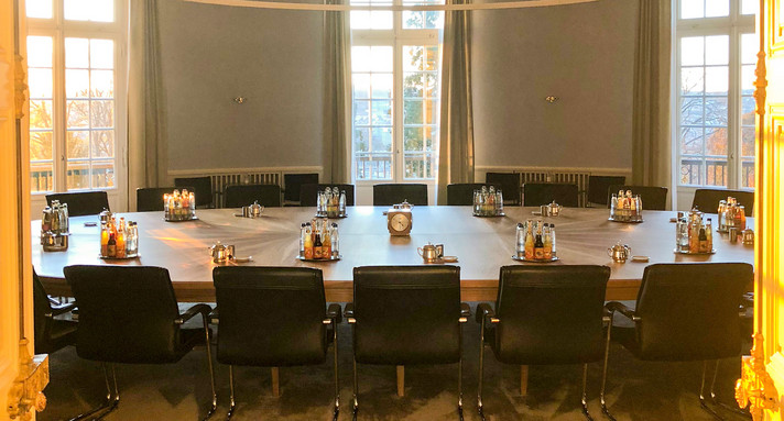 Beschreibung vom Bild: Man sieht den Raum in dem die Landesregierung tagt. Er ist in der Villa Reitzenstein. Das ist der Kabinett-Saal. Hier treffen sich die Minister und Ministerinnen zu Besprechungen. Es ist Abend. Im Raum sind nur ein großer runder Tisch, Stühle und Getränke. Die Sonne geht unter. Sie scheint in den Raum. Dadurch ist das Licht im Raum gold-gelb. 