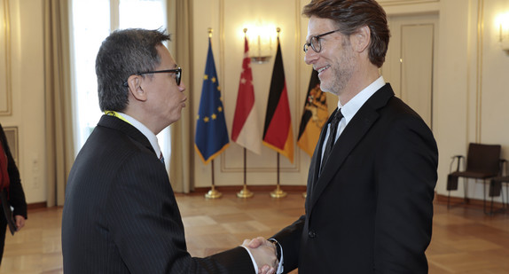 Staatsminister Dr. Florian Stegmann (rechts) begrüßt den Botschafter der Republik Singapur, Lee Chong Hock (links). Im Hintergrund stehen Fahnen.