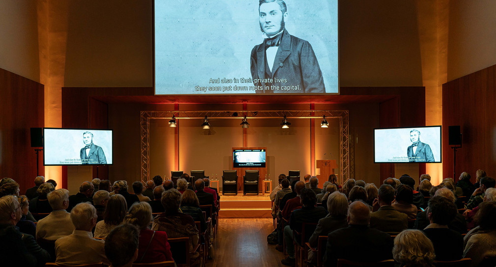 Die Veranstaltungsgäste sehen ein kurzes Video zur Biographie James Simons über die Leinwand im Großen Saal