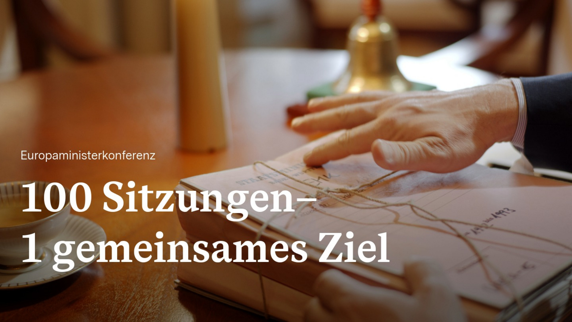 Hand in Anzug legt auf einem Tisch einen zusammengebundenen Stapel Akten Buch ab. Text auf Bild: Europaminsiterkonferenz - 100 Sitzungen, 1 gemeinsames Ziel. 