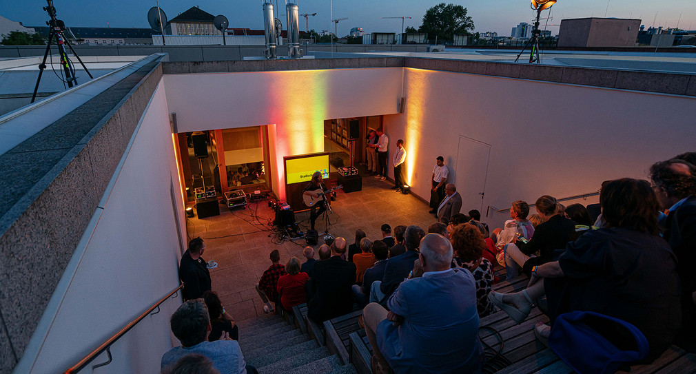 Singer-Songwriterin Anne Haigis performt auf der Dachterrasse. Die Gäste sitzen auf den Stufen der Dachterrasse und genießen den sommerlichen Abend. Im Hintergrund Skyline von Berlin.