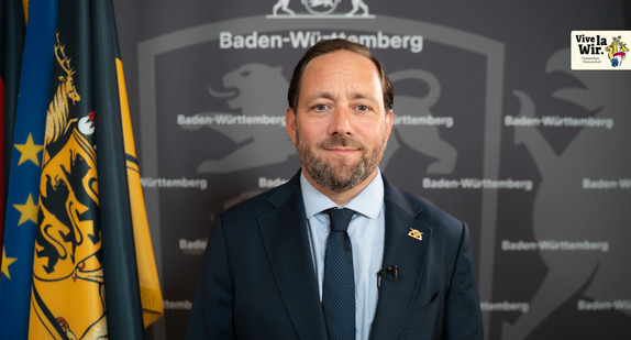 Staatssekretär Florian Haßler vor einer grauen Wand, auf der "Baden-Württemberg" steht. Hinter ihm sind die europäische, die deutsche und die baden-württembergische Flagge zu sehen.