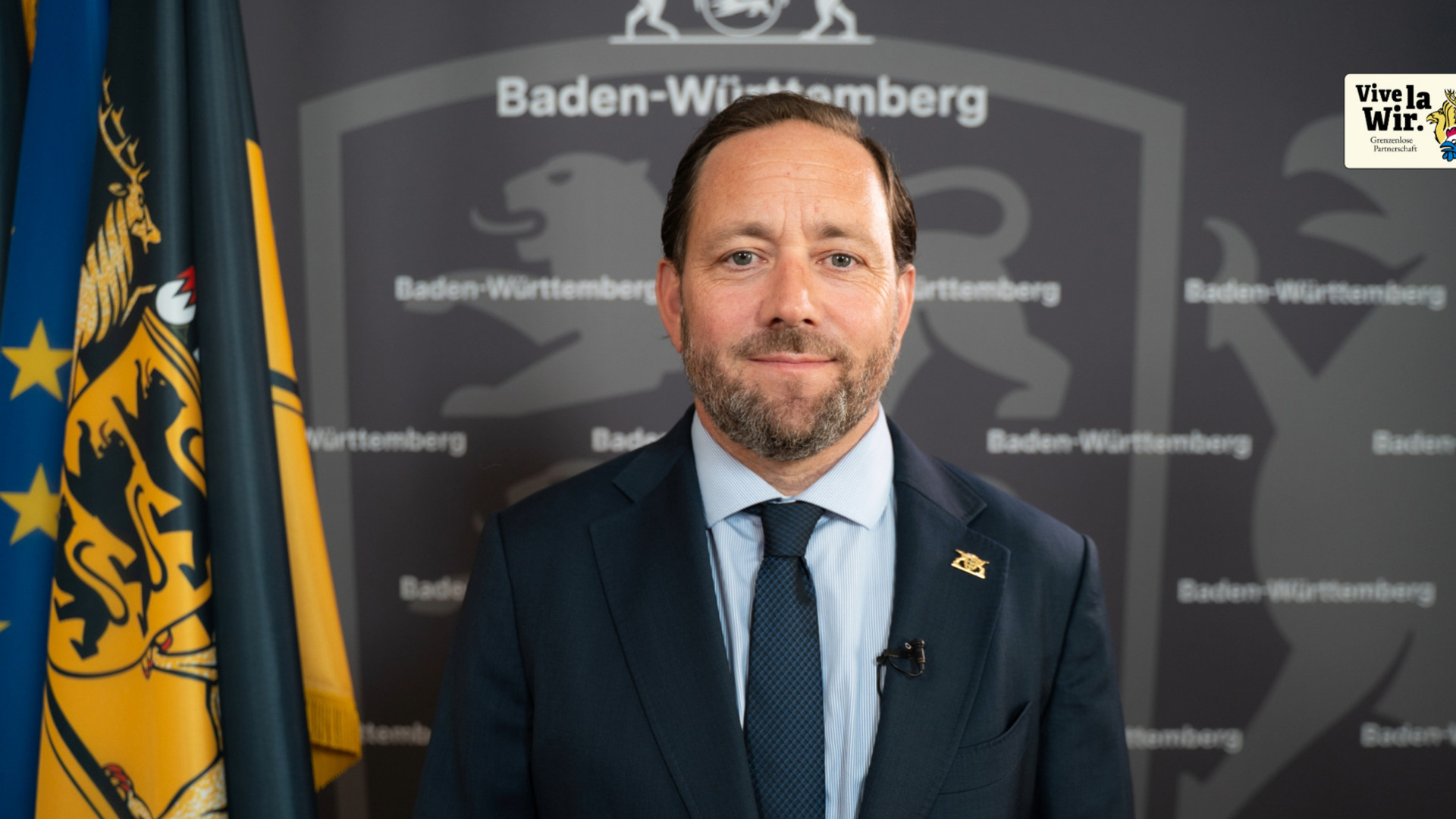 Staatssekretär Florian Haßler vor einer grauen Wand, auf der "Baden-Württemberg" steht. Hinter ihm sind die europäische, die deutsche und die baden-württembergische Flagge zu sehen.