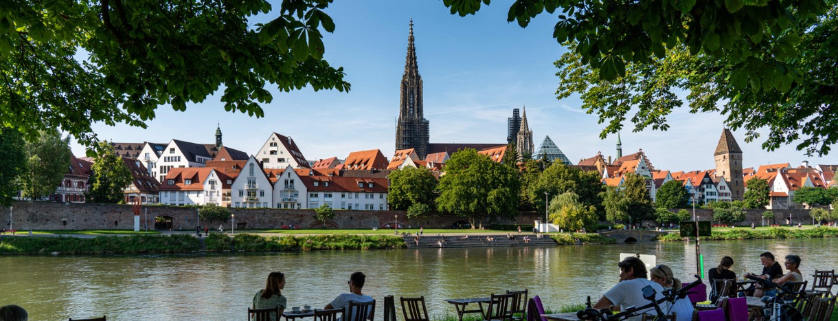 Menschen sitzen in einem Biergarten am Donauufer mit Blick auf das Ulmer Münster und die Altstadt.