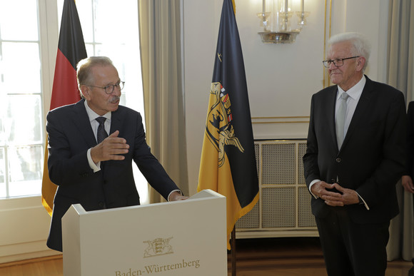 Franz Fehrenbach (links) und Ministerpräsident Winfried Kretschmann (rechts) stehen stehen im Runden Saal der Villa Reitzenstein vor Fahnen.