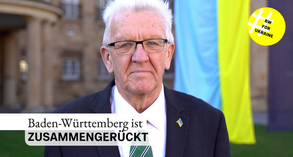 Ministerpräsident Winfried Kretschmann steht bei einer Ansprache zum Jahrestag des russischen Angriffskrieges gegen die Ukraine vor der Villa Reitzenstein in Stuttgart. Im Hintergrund ist die ukrainische Flagge zu sehen.