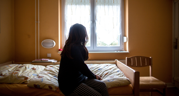 Eine Frau sitzt zum Fenster blickend in einem Frauenhaus auf einem Bett.
