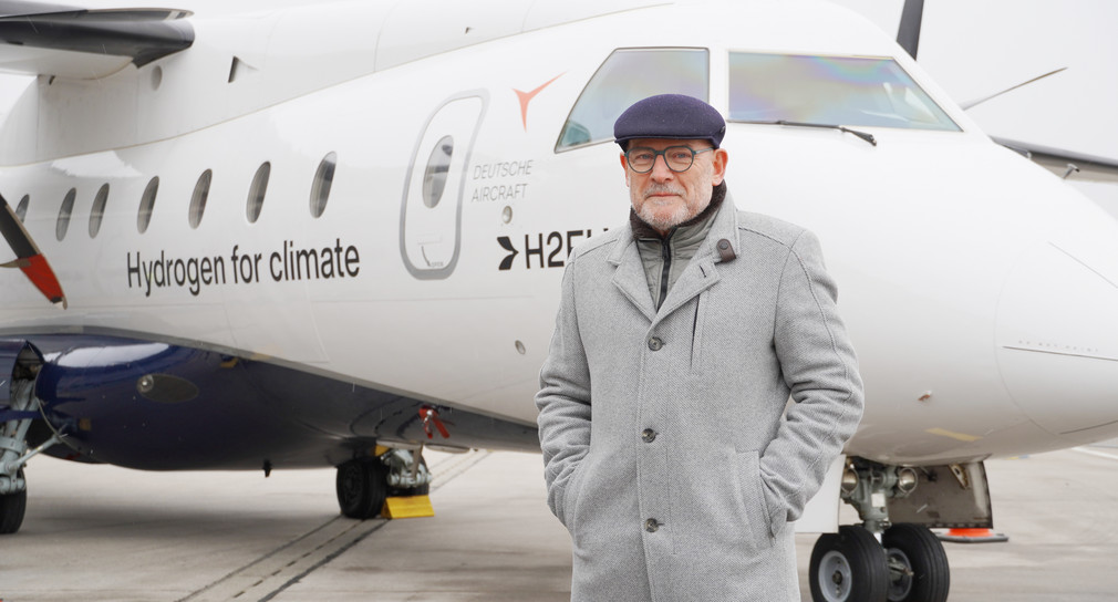 Minister Hermann steht vor dem Flugzeug Hangar. Er trägt eine Mütze und einen grauen Mantel.