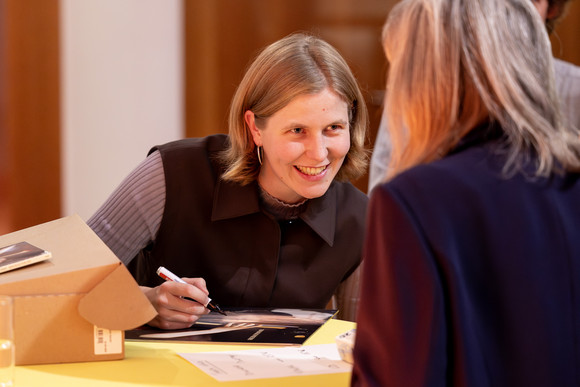 Tabea Kind gibt ein Autogramm und beug sich lachend in Richtung Kamera zu Besucherin, die man nur von hinten sieht.