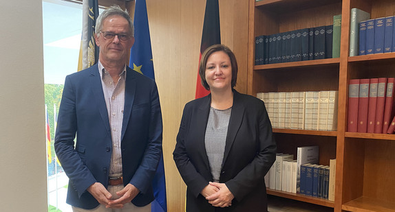 Staatssekretär Rudi Hoogvliet steht zusammen mit Frau Ana Polak Petrič, Botschafterin der Republik Slowenien im Kaminzimmer der Landesvertretung. Im Hintergrund ist ein Bücherregal zu sehen und von links nach recht die Baden-Württemberg-Flagge, die Europa-Fahne und die Deutschland-Flagge.