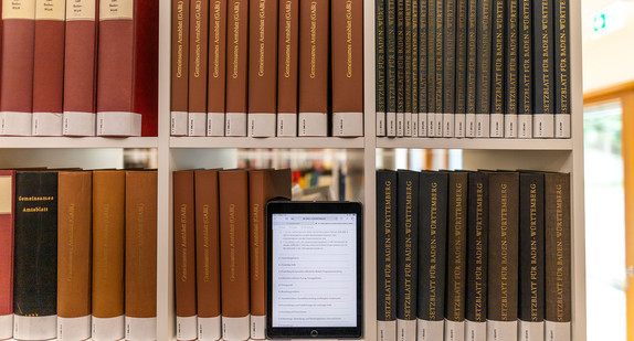 In einer Bücherei stehen Gesetzesblätter des Landes Baden-Württemberg und ein Tablet mit einem Gesetzestext.