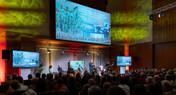 Blick in den großen Saal der Landesvertretung. Links sitzen die vier Panelteilnehmerinnen und Teilnehmer. Rechts sieht man die Band. Im Vordergrund das zahlreiche Publikum.