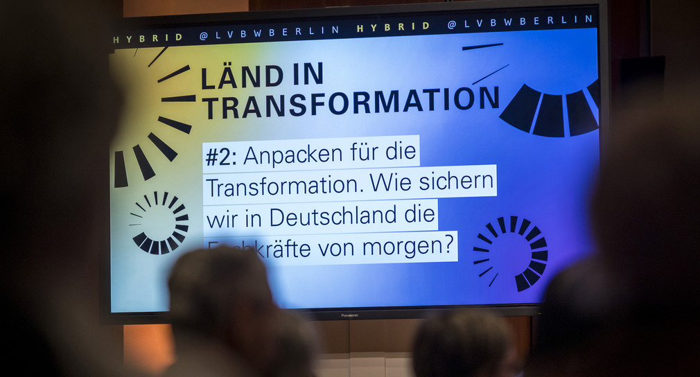 Auf einem Bildschirm steht das Veranstaltungsmotto und das Thema des Abends: LÄND IN TRANSFORMATION - Anpacken für die Transformation. Wie sichern wir in Deutschland die Fachkräfte von morgen?