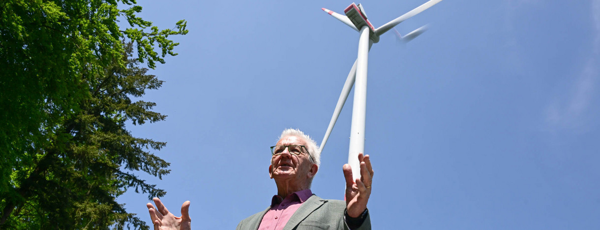 Ministerpräsident Winfried Kretschmann steht bei einer Besichtigung einer Baustelle einer Windkraftanlage vor einem schon gebauten Windrad.