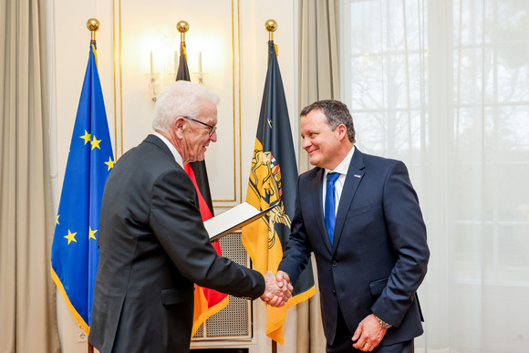 Ministerpräsident Winfried Kretschmann (links) überreicht Thomas Speidel (rechts) die Staufermedaille in Gold. Im Hintergrund stehen Fahnen.