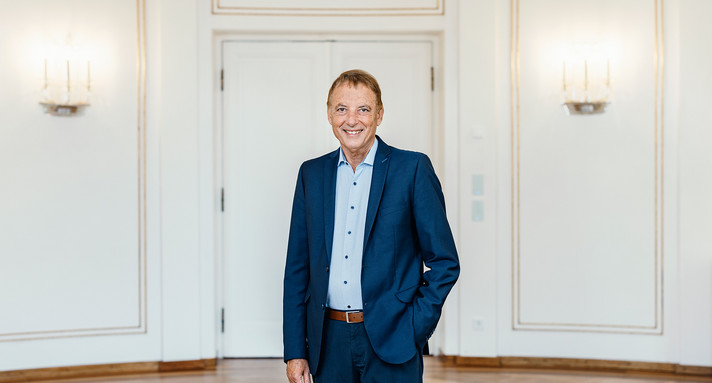 Staatsminister Jörg Krauss steht in blauem Anzug und weißem Hemd mit einer Hand in der Hosentasche vor weißen Doppeltüren in einem eleganten Raum. 