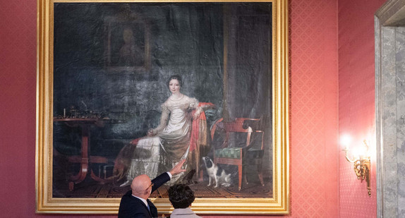 Ein Besucher und eine Besucherin einer Veranstaltung zur Enthüllung des Gemäldes „Königin Catharina von Württemberg in ihrem Arbeitszimmer im Neuen Schloss“ stehen im Empire-Zimmer des Neuen Schlosses in Stuttgart vor dem Gemälde.