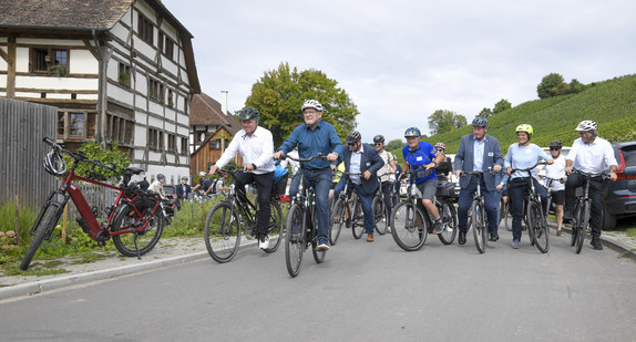 Minister steht mit einer Gruppe Fahrradfahrer auf einem Radweg am Bodensee. Er trägt einen Helm. 