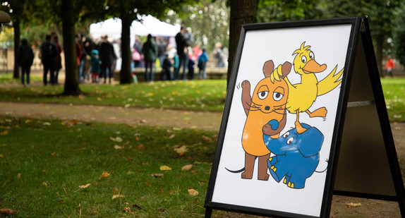 Plakat-Aufsteller mit den Figuren Maus, Elefant und Ente aus der Sendung mit der Maus auf einer Wiese mit Bäumen und Menschen im Hintergrund.
