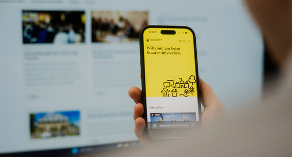Man sieht eine Hand. Die Hand hält ein Smartphone. Auf dem Smartphone sieht man die Internet Seite Staatsministeriums Baden-Württemberg. 