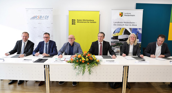 Eine Gruppe von Menschen sitzt am Tisch und schaut in die Kamera. Vor ihnen liegen Dokumente zum unterschreiben. 