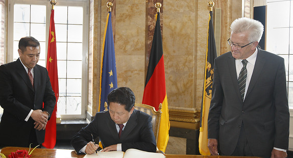 Gouverneur Qiufa Chen (M.) trägt sich in das Gästebuch der Landesregierung ein. Daneben steht Ministerpräsident Winfried Kretschmann (r.)