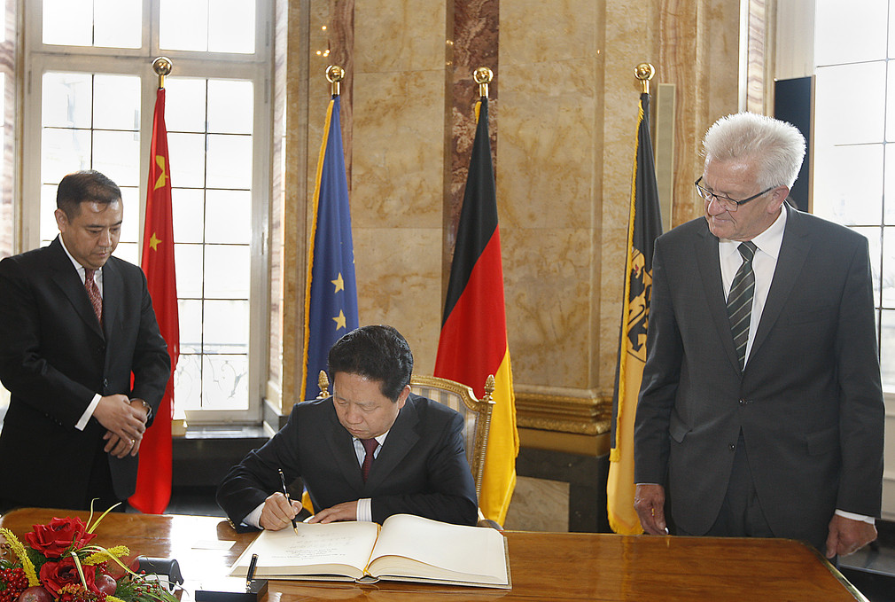 Gouverneur Qiufa Chen (M.) trägt sich in das Gästebuch der Landesregierung ein. Daneben steht Ministerpräsident Winfried Kretschmann (r.)