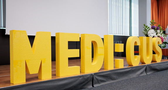 Schriftzug „MEDI:CUS“ auf einer Bühne