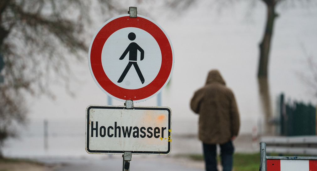 Ein Mann geht hinter einem Schild mit der Warnung „Hochwasser“ zu einem Flussufer.