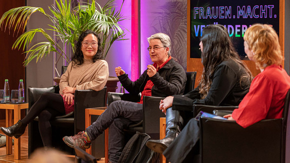 Drei Frauen auf dem Podium, links Yumin Li, rechts daneben sitzt Lourdes Martínez und spricht in das Mikrophon. Nur von hinten zu sehen: Mina Saidze und Dörthe Eickelberg