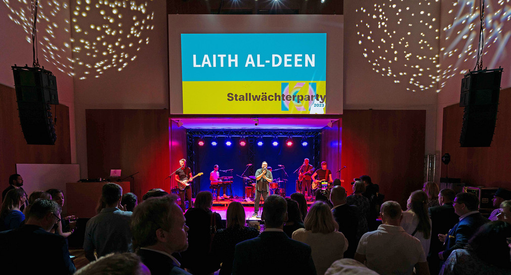 Der gebürtige Karlsruher Laith Al-Deen steht auf der Bühne im Großen Saal. Stimmungsvolle Beleuchtung und auf der Leinwand über der Bühne steht "Laith Al-Deen und Stallwächterparty 2023".