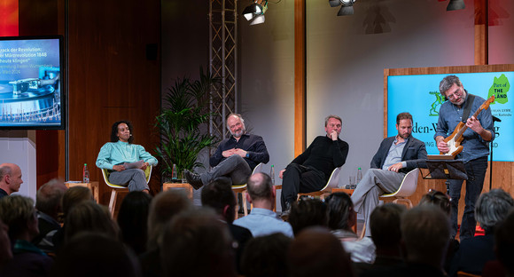 Die Panelteilnehmerinnen und -teilnehmer und das Publikum hören einer Interpretation von Jo Ambros und seiner Band zu