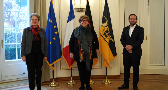 von links nach rechts: Staatsrätin Barbara Bosch, Brigitte Torloting, Vizepräsidentin der Region Grand Est, und Staatssekretär Florian Hassler stehen vor Fahnen.