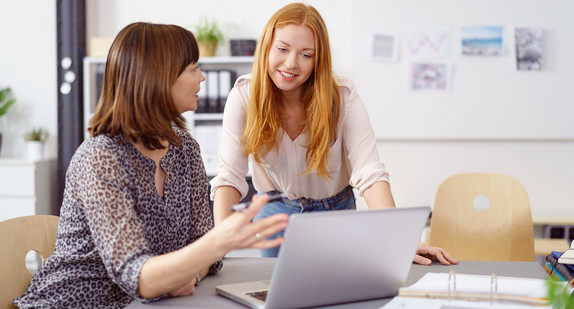 In einem Büro unterhalten sich zwei Frauen. Eine sitzt an einem Tisch und eine steht neben dem Tisch. Auf dem Tisch steht ein Laptop.