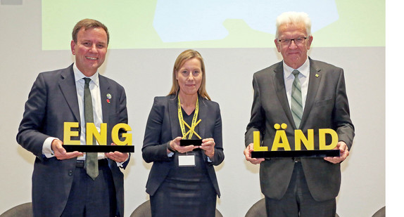 Baden-Württembergs Ministerpräsident Winfried Kretschmann (rechts) steht bei der Eröffnung der neuen Vertretung von Baden-Württemberg in London neben dem britischen Minister für Wirtschaft und Energie, Greg Hands (links), und Nicola Pinder (Mitte), Vertreterin von Baden-Württemberg in Großbritannien, auf der Bühne. Sie halten einen Schriftzug „ENG X LÄND“ in den Händen.