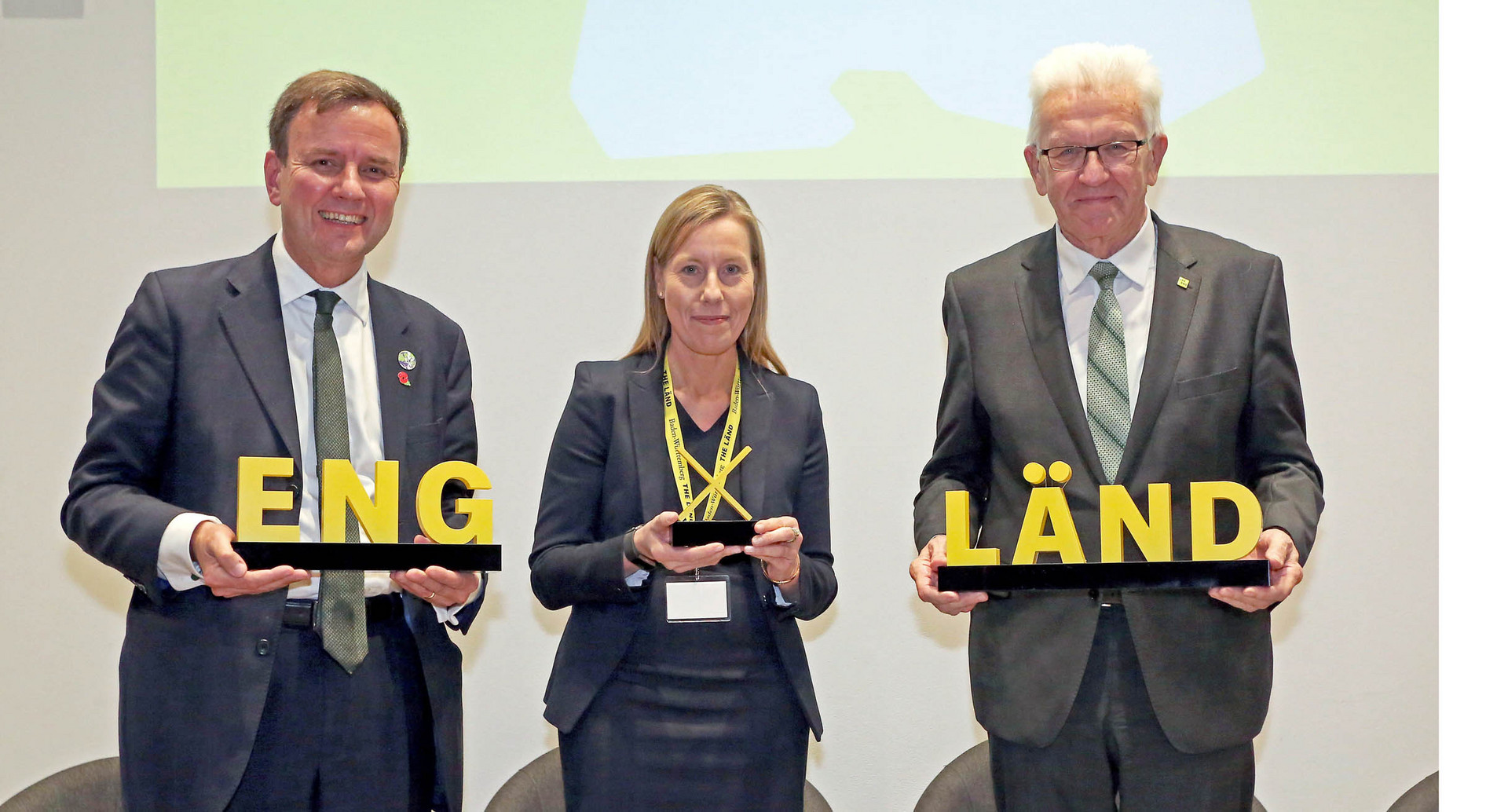 Baden-Württembergs Ministerpräsident Winfried Kretschmann (rechts) steht bei der Eröffnung der neuen Vertretung von Baden-Württemberg in London neben dem britischen Minister für Wirtschaft und Energie, Greg Hands (links), und Nicola Pinder (Mitte), Vertreterin von Baden-Württemberg in Großbritannien, auf der Bühne. Sie halten einen Schriftzug „ENG X LÄND“ in den Händen.