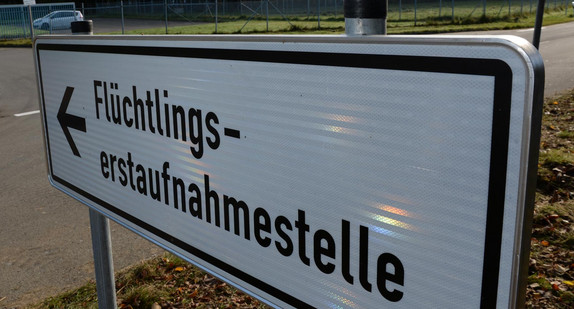 Ein Schild mit der Aufschrift „Flüchtlingserstaufnahmestelle“, im Hintergrund ein Fahrzeug an einer Einfahrt.