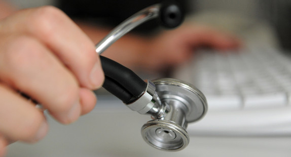 Ein Arzt hält in einem Behandlungszimmer in seiner Praxis in Deizisau im Landkreis Esslingen ein Stethoskop in der Hand, mit der anderen Hand bedient er eine Computertastatur. (Bild: dpa)