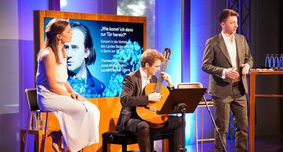 Auf der Bühne sitzt die Sopranistin Theresa Pilsl auf einem Barhocker und lauscht dem Gesang Bariton Jonas Müller. Zwischen beiden sitzt Gitarrist Leonard Becker und begleitet.