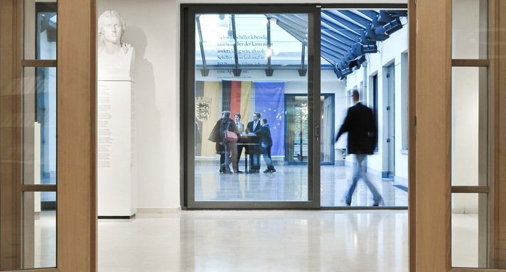 Eingangssituation mit Blick in den glasüberdachten Innenhof mit Gruppe von Menschen vor Fahnen.