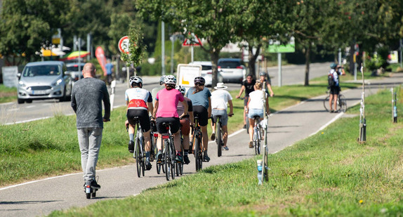 Zahlreiche Menschen sind auf einem Radweg bei Sonnenschein auf ihren Fahrrädern und E-Roller unterwegs.